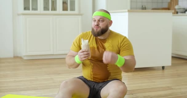Sweaty fat man in yellow sportswear eating danar sitting on fitness mat at home. Royalty Free Stock Footage
