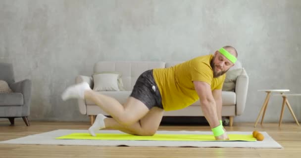 Man in yellow sportswear doing legs lifting exercises on mat on knees at home. — Stock Video