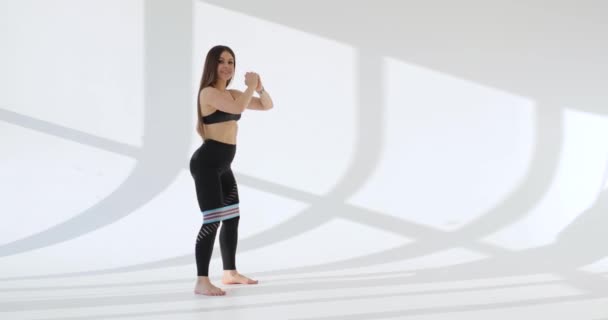Sett fra siden av unge, hvite kvinner som trener på knebøy i hvit, solrik bakgrunn. – stockvideo