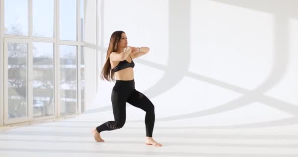 Vista lateral de la joven mujer caucásica haciendo ejercicios en un estudio blanco con una gran ventana en el fondo . — Vídeo de stock