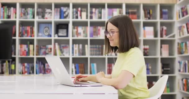 젊은 여성 이 책상에 앉아 노트북을 들고 도서관에서 일하면서 웃고 있습니다. 그녀는 인터넷을 통해 소통 합니다. 사이드 뷰. 온라인 컨퍼런스. — 비디오