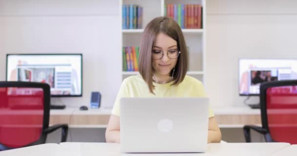 La joven está sonriendo, sentada en un escritorio con un portátil, trabajando en la biblioteca. Se comunica a través de Internet. Vista frontal. Conferencia en línea. — Vídeos de Stock