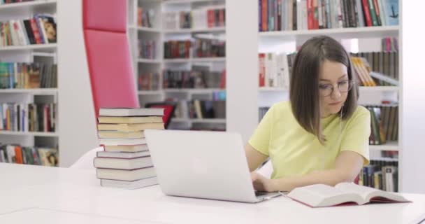 年轻的女人坐在书桌前，手里拿着笔记本电脑，在图书馆里工作。前景. — 图库视频影像