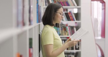 Gözlüklü güzel Avrupalı kız öğrenci üniversite kütüphanesinde kitap okuyor. Yan görünüm.