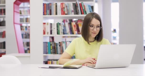 Молодая женщина улыбается, сидит за столом с ноутбуком, работает в библиотеке. Она общается через Интернет. Вид спереди. Онлайн-конференция. — стоковое видео