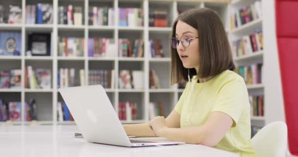 Jovem estudante está sentado em uma mesa com um laptop, trabalhando na biblioteca. Ela se comunica através da Internet. Vista lateral. No fundo é um rack com livros. Conceito de ensino à distância. — Vídeo de Stock