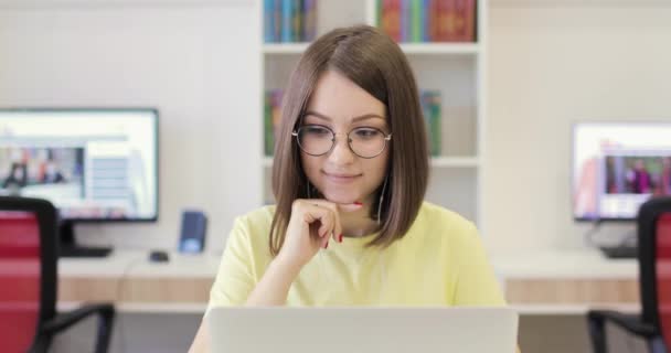 Unga kvinna ler, sitter vid ett skrivbord med en bärbar dator, arbetar i biblioteket. Hon kommunicerar via internet. Framifrån. Nätkonferens. — Stockvideo