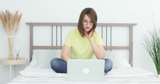 Jovem mulher bonita está sentada na cama em frente a um laptop. Ela está se comunicando via link de vídeo. Vista frontal. — Vídeo de Stock