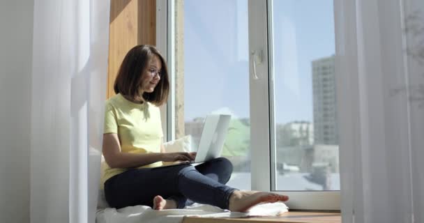 Genç ve güzel bir kadın pencere eşiğinde oturuyor ve bir dizüstü bilgisayar kullanarak sohbet ediyor. Yan görünüm. — Stok video