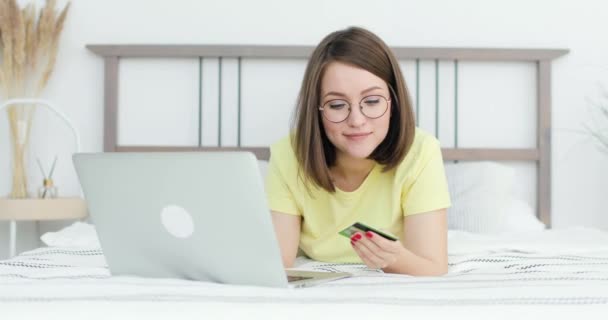 Jeune belle femme est allongée sur le lit devant un ordinateur portable. Elle effectue un paiement en ligne. Vue de face. — Video
