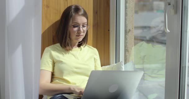 Jeune belle femme avec ordinateur portable est assis près de la fenêtre et bavarder. Vue de face. — Video