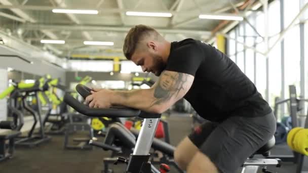 Atleta adulto maschio con tatuaggi sul braccio sta facendo esercizi su un simulatore di ciclismo. — Video Stock