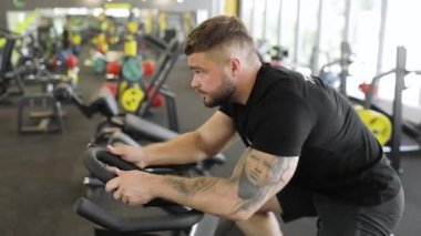 Siyah tişörtlü kolunda dövmeleri olan yetişkin bir erkek sporcu bisiklet simülatörü üzerinde egzersiz yapıyor. Yan görünüm.