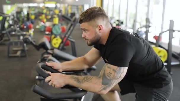 Dospělý sportovec s tetováním na paži v černém tričku cvičí na cyklistickém simulátoru. Boční pohled. — Stock video