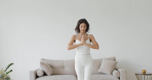 Ασκήσεις γιόγκα, μαθήματα στο σπίτι. Μπροστά. Μια νεαρή γυναίκα με λευκό μπλουζάκι διατηρεί την ισορροπία ενώ στέκεται στο ένα πόδι.. — Αρχείο Βίντεο
