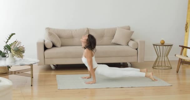 O femeie tânără într-un vârf alb face exerciții de yoga pe covor într-o cameră de zi ușoară. Vedere laterală . — Videoclip de stoc
