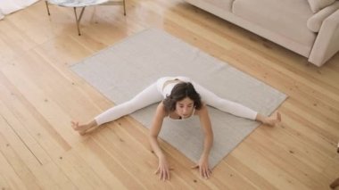 Beyaz elbiseli genç bir kadın oturma odasındaki bir paspasın üzerinde esneme egzersizleri yapıyor. Üst görünüm.