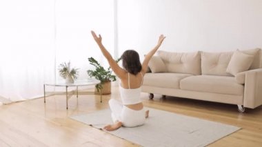 Beyaz elbiseli genç bir kadın pencerenin önündeki parlak oturma odasında minder üzerinde yoga egzersizleri yapıyor. Arka plan.