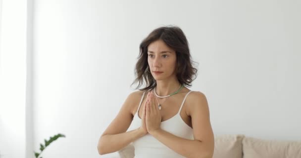 Una hermosa morena en un top blanco está haciendo ejercicios de yoga en casa en una luminosa sala de estar y meditando. Primer plano. — Vídeos de Stock