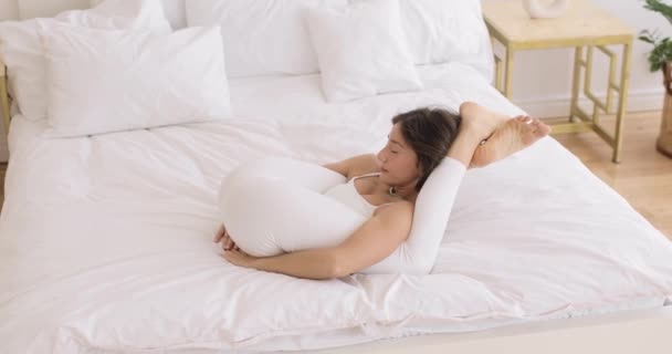 Morena flexible en dormitorio blanco haciendo ejercicios de estiramiento con la pierna detrás de la cabeza. Meditación. Vista lateral. — Vídeo de stock