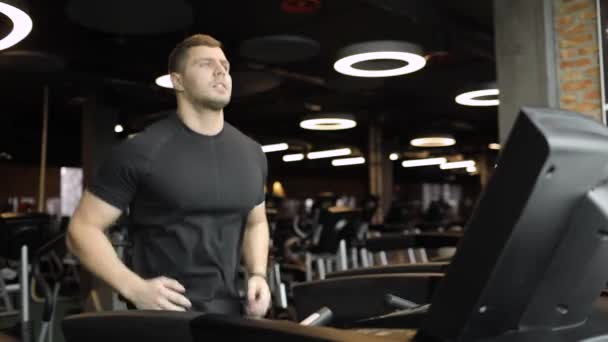 Siyah tişörtlü atletik genç adam spor kulübünde modern koşu bandı üzerinde kardiyo egzersizi yapıyor, gülümsüyor.. — Stok video