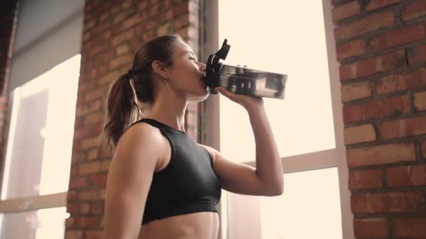 Retrato de mujer en forma hermosa en un tanque negro corto beber agua en un gimnasio, después de un intenso entrenamiento, sonriendo — Vídeos de Stock