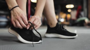 Eğitim için hazırlık. Spor ayakkabılarını içeride bağlayan genç bir kadının bacaklarına yakın çekim. Arka planda, bir adam şınav çekiyor..