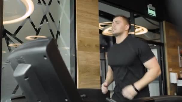 Спортивный юноша в черной футболке делает кардио-упражнения на современной беговой дорожке в спортклубе. — стоковое видео