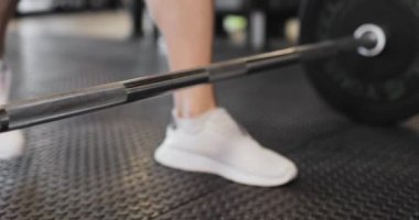 Sporcular bara yaklaşıyorlar. Beyaz spor ayakkabılı haltercinin kısmi görüntüsü halter kaldırıyor.