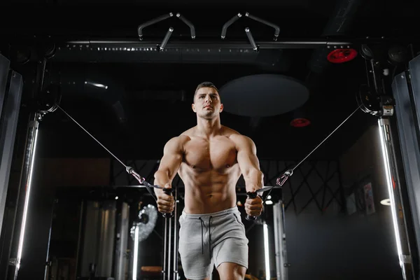 Type bodybuilder modèle avec un torse nu, trains dans la salle de gym. Les muscles sont tendus. Fond sombre. — Photo