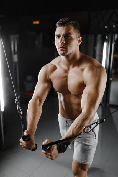 Type bodybuilder modèle avec un torse nu, trains dans la salle de gym. Les muscles sont tendus. Fond sombre. — Photo