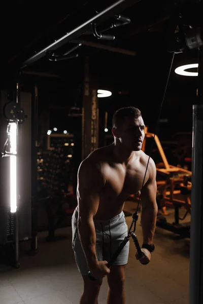 Type bodybuilder modèle avec un torse nu, trains dans la salle de gym. Les muscles sont tendus. Fond sombre. — Photo