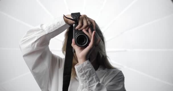 Retrato de una joven fotógrafa con estilo en una camisa blanca con una nariz perforada y el pelo largo y rubio, haciendo fotos, mirando a las cámaras de lente de vídeo y sonriendo. — Vídeos de Stock