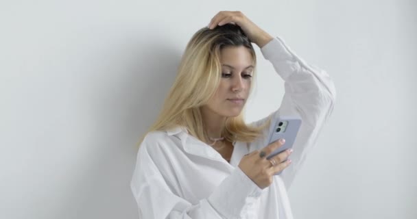 Ritratto di giovane donna elegante in camicia bianca con capelli biondi, su sfondo bianco in studio. Utilizzando un telefono cellulare, digitando, sfogliando le pagine. — Video Stock