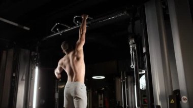 Çıplak gövdeli yakışıklı genç sporcu spor salonundaki yatay çubukta kaslarını şişiriyor ve barfiks çekiyor. Arkadan bak. Etkin yaşam biçimi.