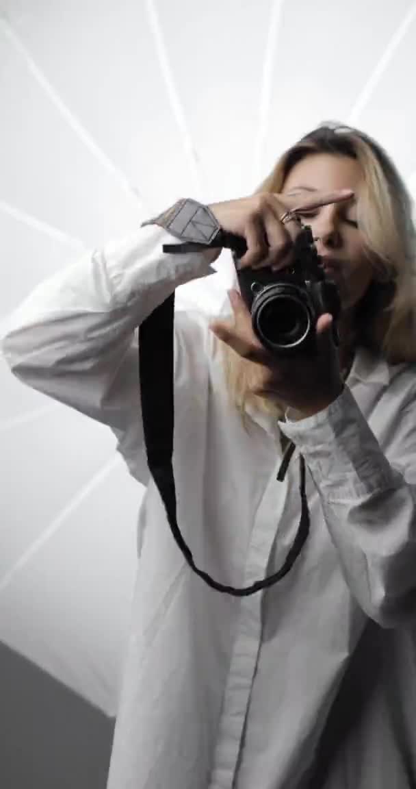 Portret van een mooie jonge fotografe in een wit shirt met lang blond haar, die opnamen maakt, in de cameralens kijkt en glimlacht. Verticale video. — Stockvideo