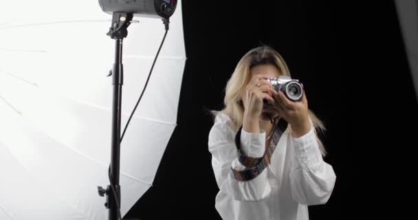 La joven fotógrafa en una camisa blanca con el pelo largo y rubio trabaja con entusiasmo haciendo fotos, utilizando equipos de iluminación. Vista lateral. — Vídeos de Stock