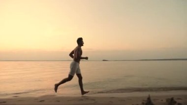 Beyaz şortlu atletik genç adam sabahın erken saatlerinde ya da gün batımında kıyıda koşuyor. Güzel gün doğumu ya da gün batımı. Yan görünüm.