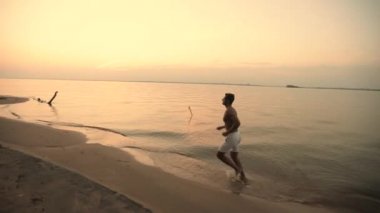 Beyaz şortlu, çıplak gövdeli atletik genç bir adam sabahları ya da gün batımında, arka planda güneşin doğuşuna ya da batışına karşı kıyıda koşuyor. Ağır çekim. Yan görünüm.