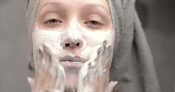 Portret van een jonge vrouw met een handdoek op haar hoofd om haar gezicht te wassen met organische schuimende reinigingsmiddelen. Het meisje reinigt de huid van het gezicht, maakt massagebewegingen. Vooraanzicht. — Stockvideo