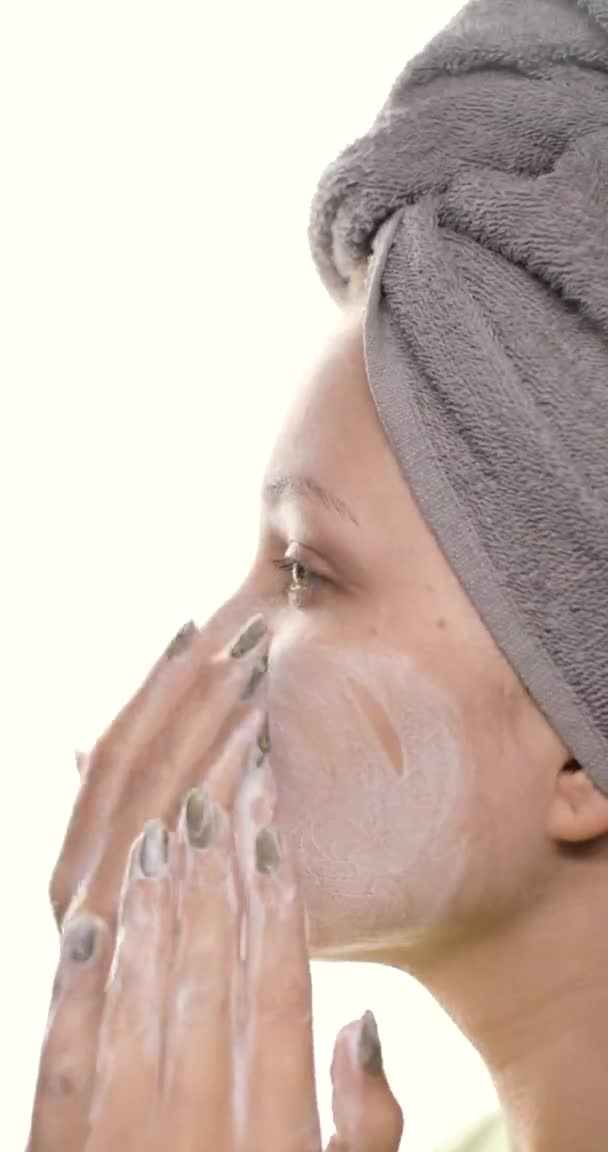 Wajah seorang wanita cantik muda dengan handuk abu-abu di kepalanya yang merawat kulitnya, membersihkan wajahnya dengan gerakan pijat menggunakan pembersih busa alami. Video vertikal. — Stok Video