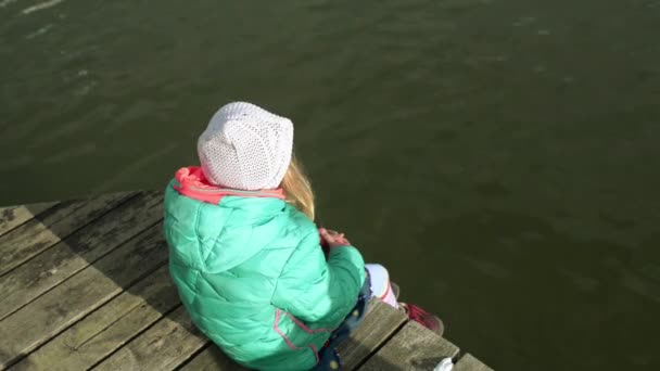 A menina bonita pequena em uma jaqueta verde senta-se em um cais de madeira em Sopot, Polônia . — Vídeo de Stock