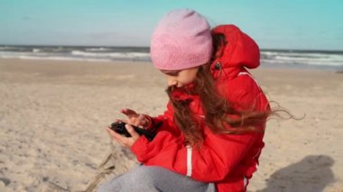 Telefon ile kırmızı ceketli güzel kız dalgaların karaya attığı odun Baltık Denizi kıyısında oturur ve mesafe içine bakıyor.