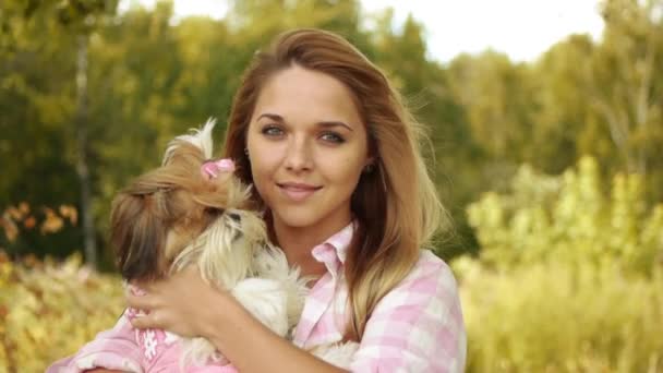 Retrato de una mujer sonriente bastante joven sosteniendo un pequeño perro esponjoso, contra el fondo del parque verde de verano — Vídeos de Stock