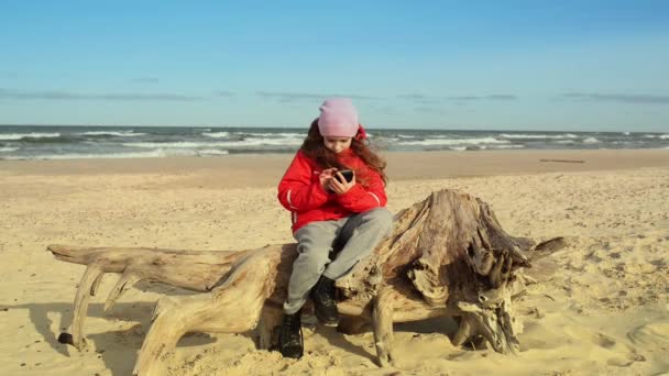 kleines hübsches Mädchen in roter Jacke mit Handy sitzt auf Treibholz am Ufer der Ostsee und blickt in die Ferne.