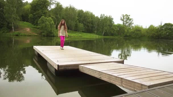 Beautiful little girl walking on a wooden bridge on a background of summer lake — Stock Video