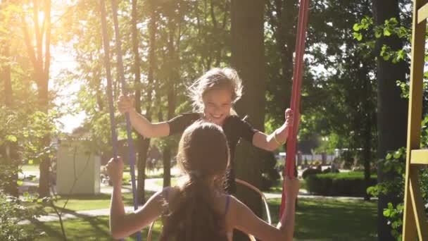 Zeitlupe: Zwei Schwestern reiten auf Schaukel im sommerlichen Stadtpark. — Stockvideo