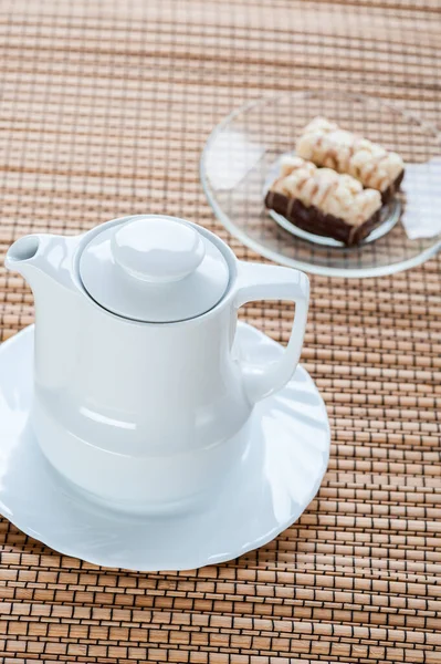 ソーサー付きの小さな白いティーポットが竹のテーブルクロスの上に立っています ミルキーチョコレートケーキ — ストック写真