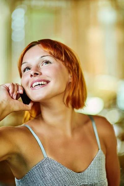 年轻的红头发女人笑着在一个有柳条椅的大厅后面用手机交谈 — 图库照片