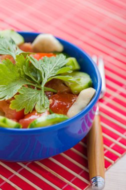 Salatalıklı salata, domates ve maydanozlu kırmızı bambu masa örtüsü üzerinde mavi bir kâse..